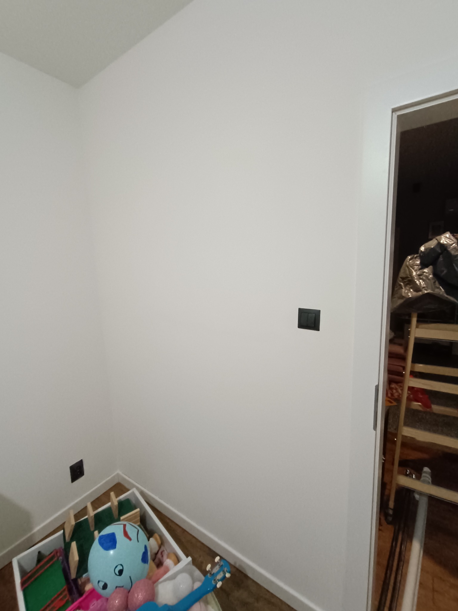 Świeżo pomalowana biała ściana w pokoju dziecięcym, widoczne gniazdka elektryczne i skrzynia z zabawkami w rogu.