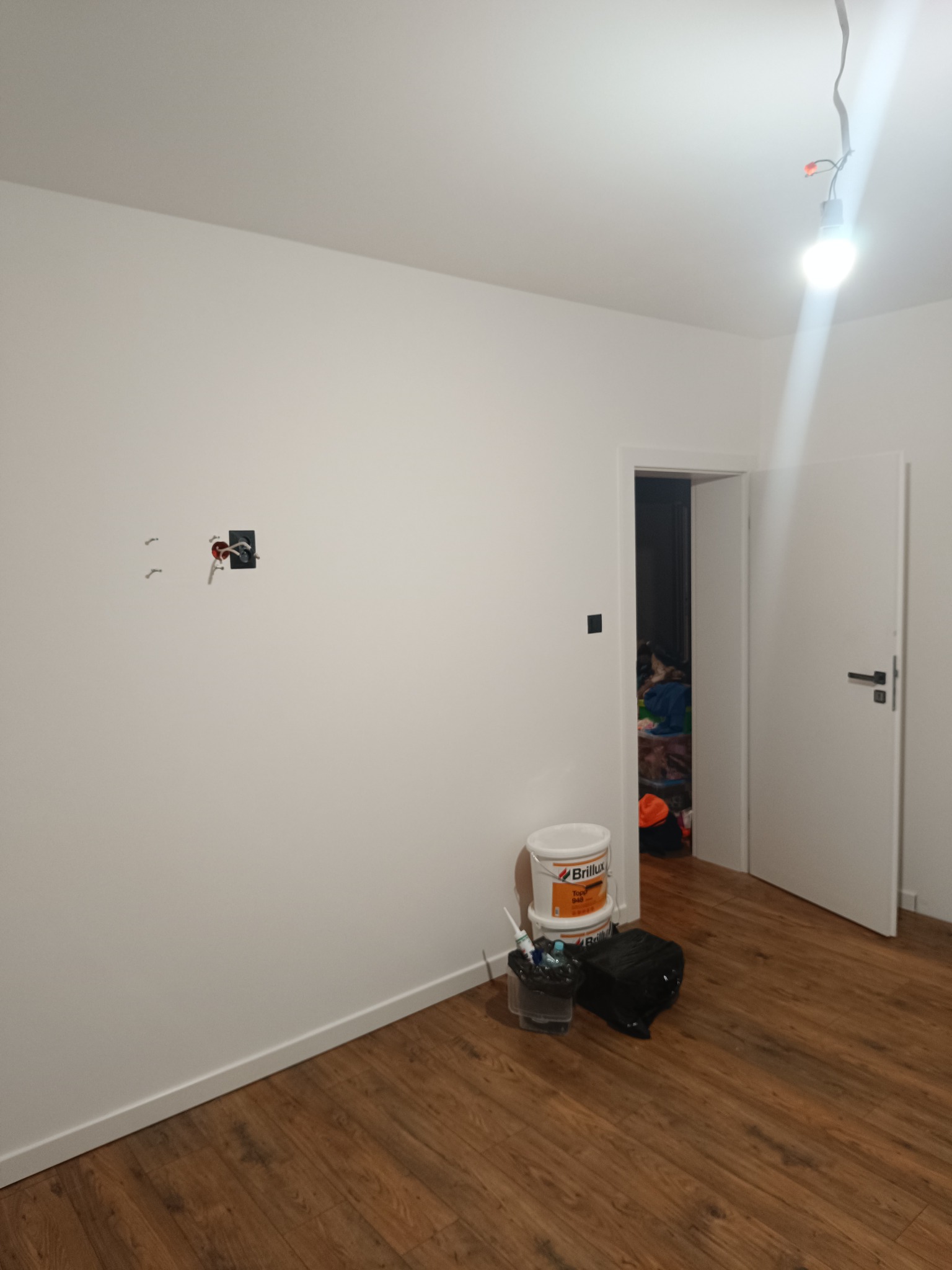 Pokój w trakcie prac wykończeniowych: ściana z puszką elektryczną bez osprzętu, wiadro z farbą Brilux, otwarte drzwi do zagraconego pomieszczenia, surowa żarówka.