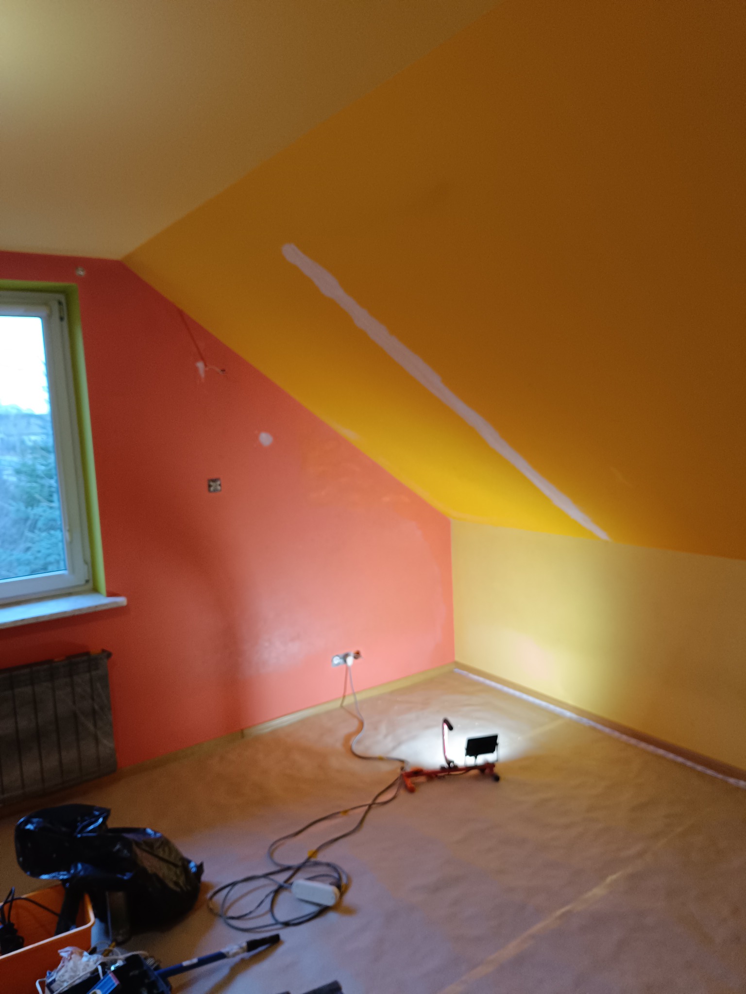 Pokój na poddaszu w trakcie odświeżania: ściany pomalowane na pomarańczowo i żółto, widoczny fragment okna, lampa halogenowa na podłodze zabezpieczonej folią malarską, oraz narzędzia malarskie w rogu.