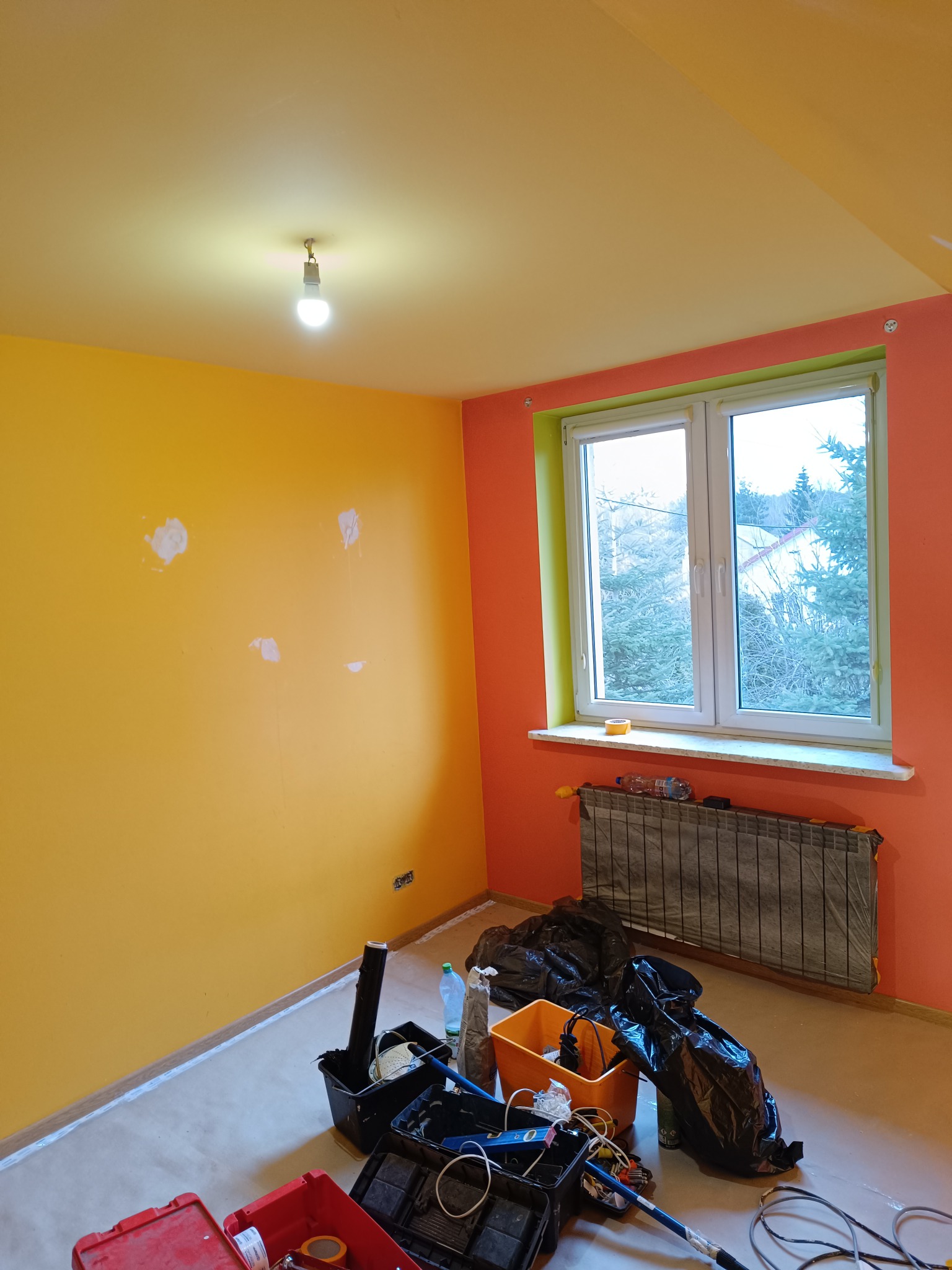 Wnętrze pokoju w trakcie malowania: żółta ściana z widocznymi miejscami po szpachlowaniu, pomarańczowa ściana przy oknie, narzędzia malarskie w kuwetach na podłodze, grzejnik zabezpieczony folią.