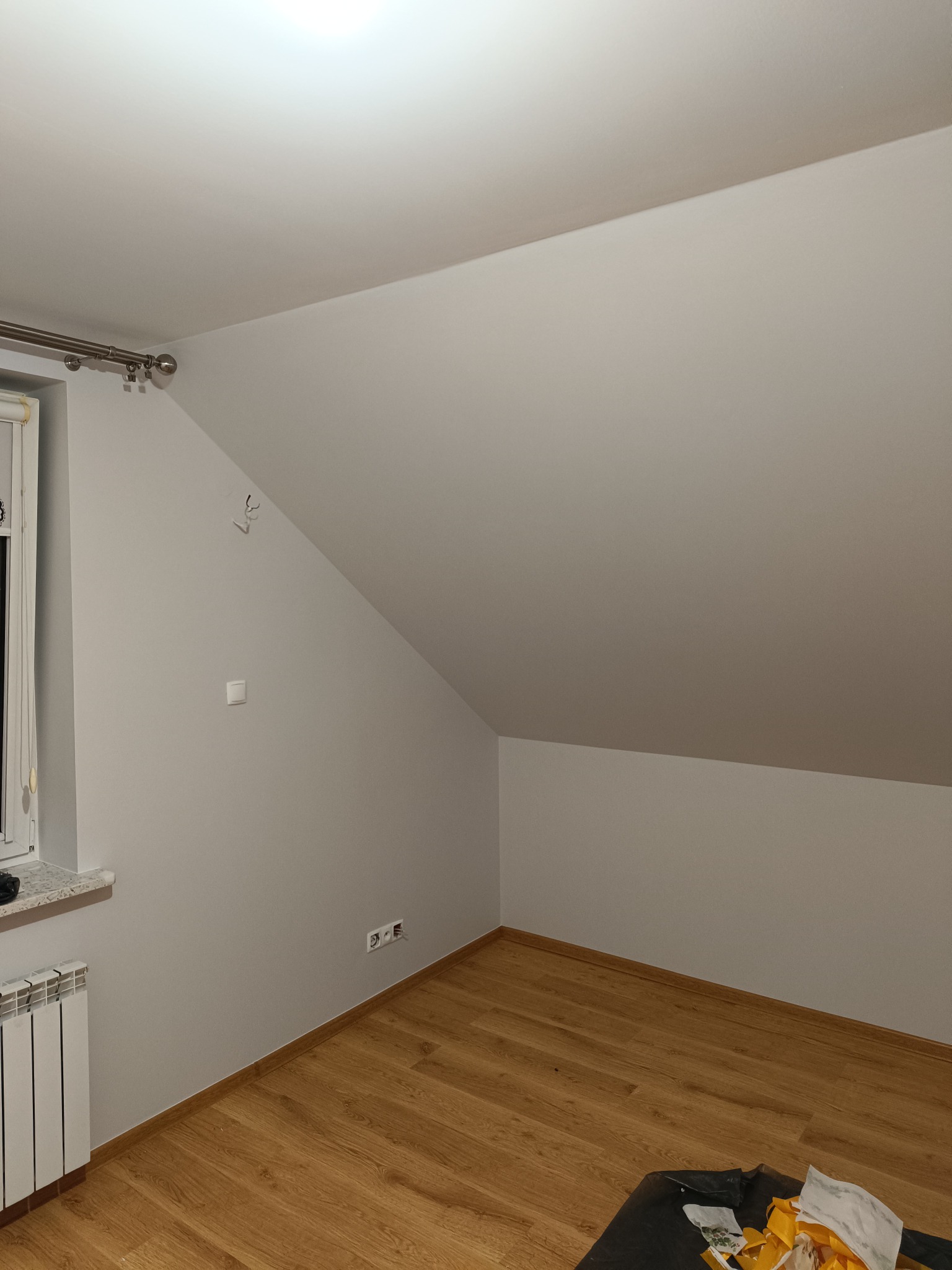 Pomalowane na szaro poddasze ze skośnym sufitem, widoczne okno z grzejnikiem, drewniana podłoga i ślady po hakach na ścianie.
