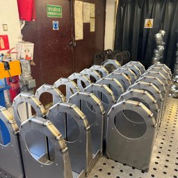 Dawid Sobczyński Usługi Spawalnicze - Szereg identycznych, spawanych konstrukcji metalowych z okrągłym otworem, ustawionych na perforowanym stole spawalniczym w warsztacie. Widoczne spoiny i elementy mocujące.