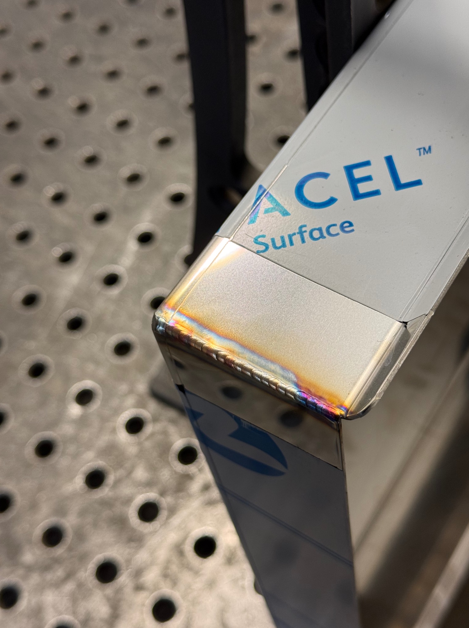 Detal spoiny TIG na krawędzi profilu z logo 'ACEL Surface', widoczne tęczowe przebarwienia termiczne na stali nierdzewnej, tło stanowi stół spawalniczy z otworami.