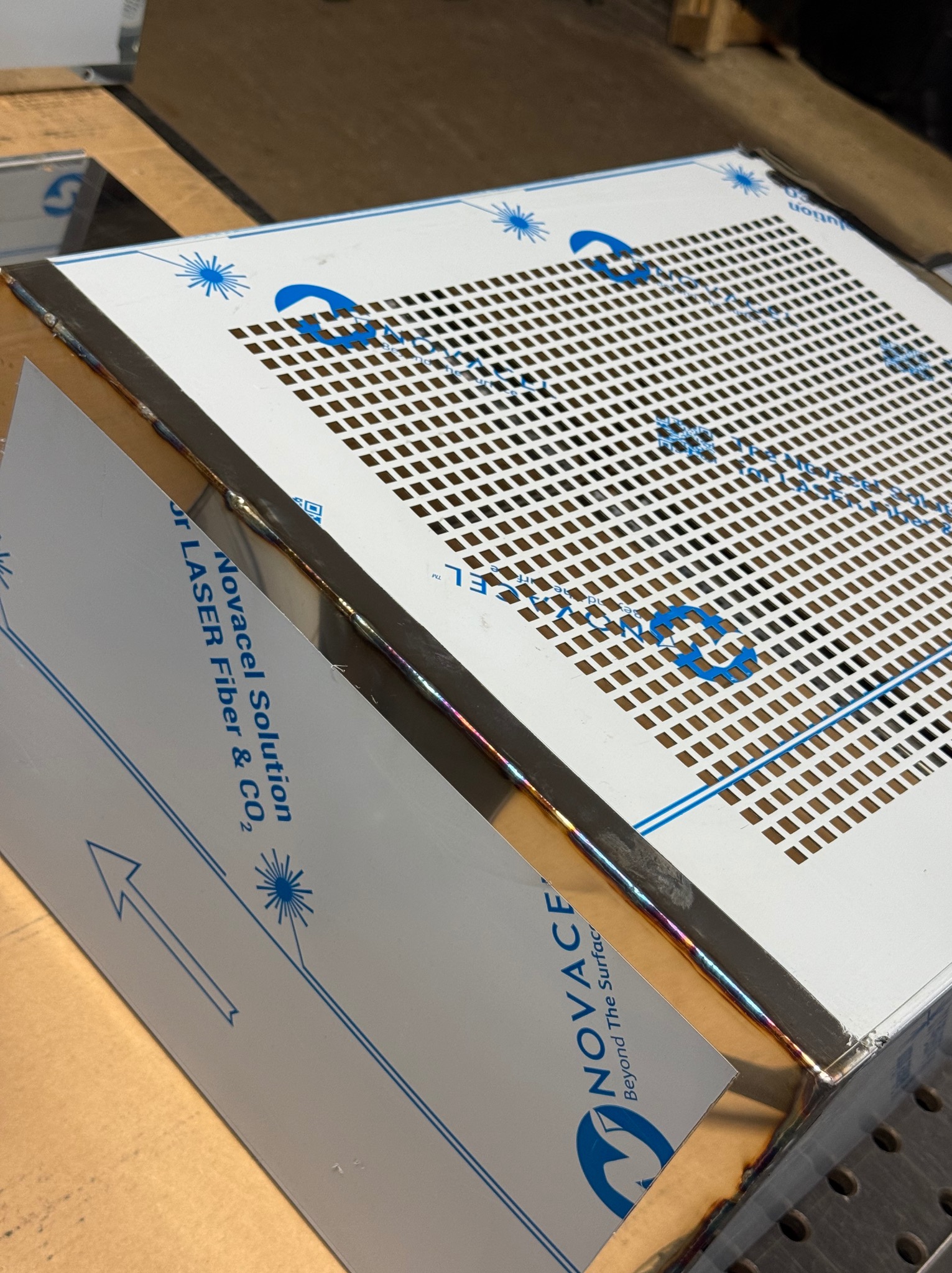 Spawana konstrukcja z perforowanej blachy nierdzewnej, widoczne ślady po spawaniu i niebieskie logo ochronnej folii Novacel.