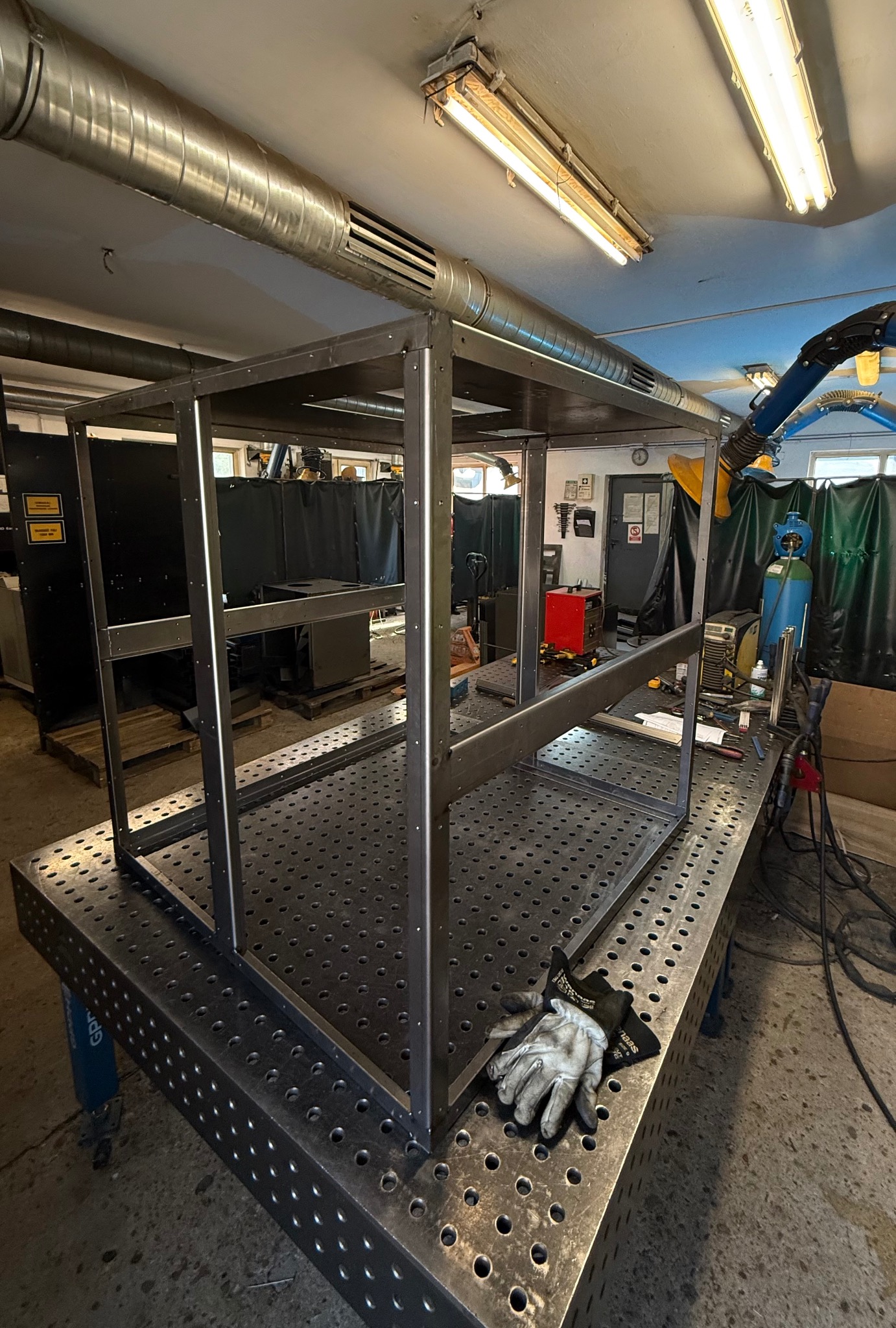 Spawana metalowa konstrukcja szkieletowa na perforowanym stole spawalniczym w warsztacie, obok leżą rękawice robocze. Widoczne przewody wentylacyjne pod sufitem.