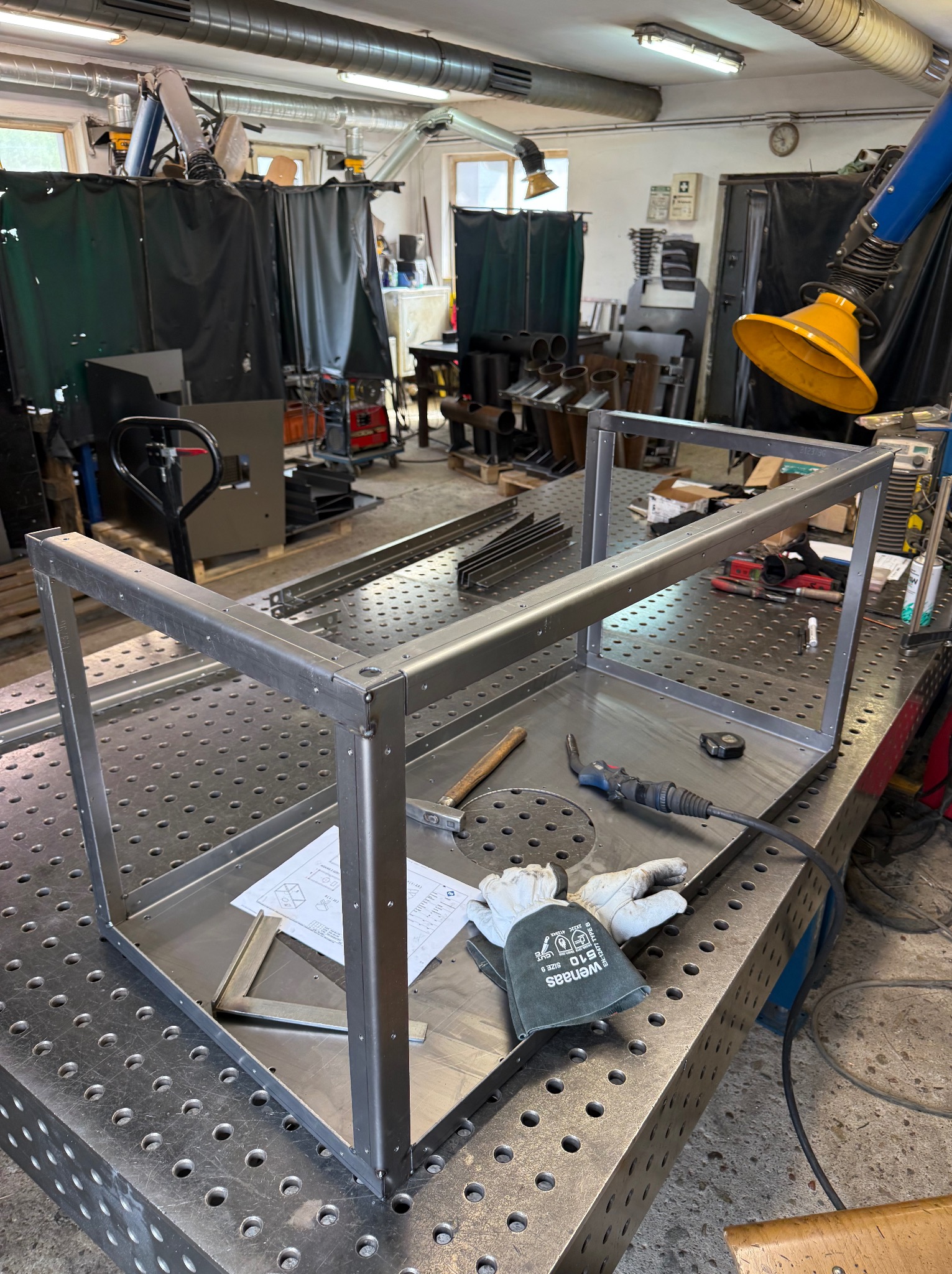 Stalowa konstrukcja spawana na perforowanym stole warsztatowym z narzędziami, rękawicami i rysunkiem technicznym. Widoczne spawy punktowe.