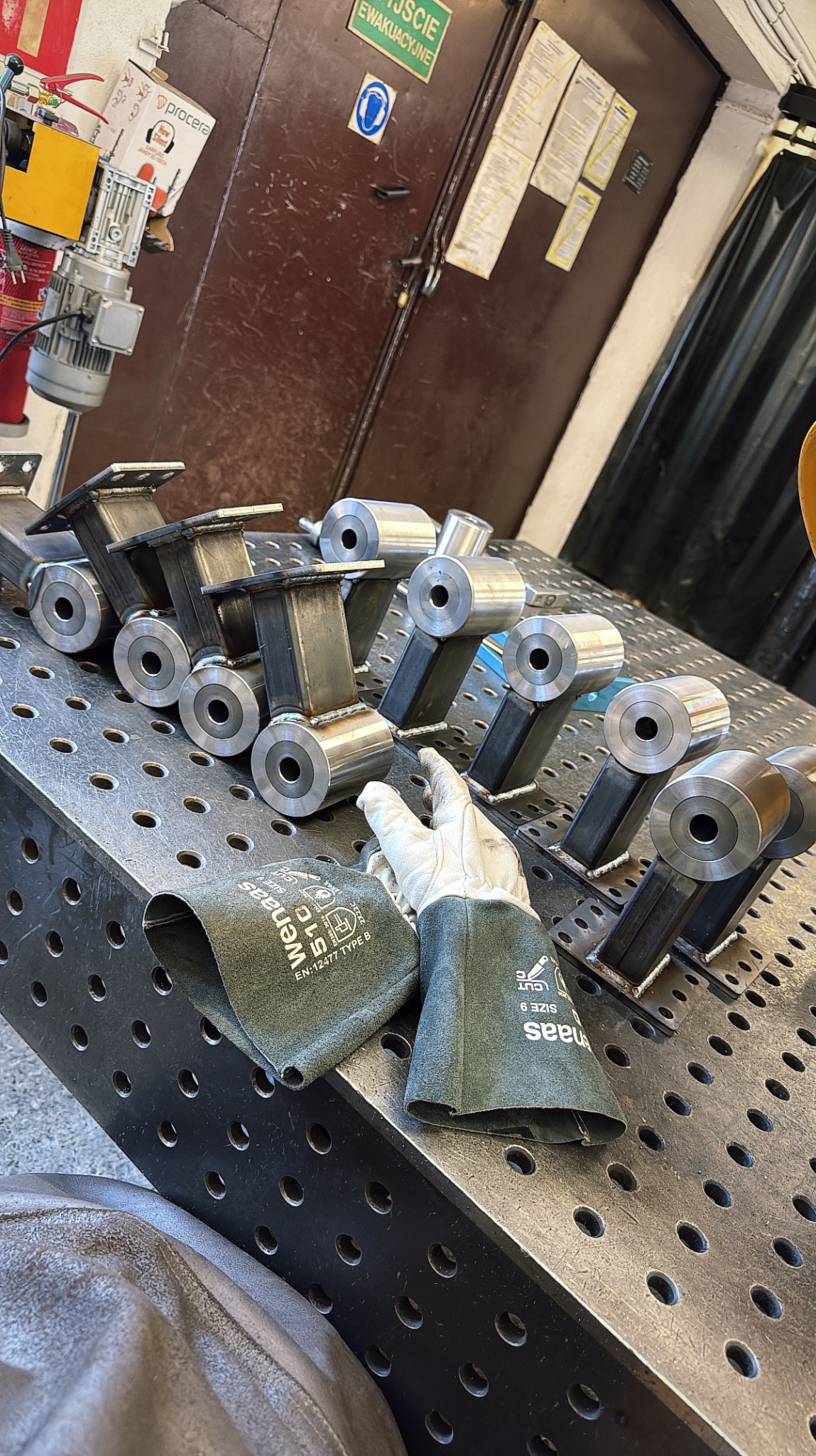 Metalowe elementy konstrukcyjne, spawane, ułożone na perforowanym stole spawalniczym, obok rękawice ochronne. Widoczny sprzęt spawalniczy w tle.