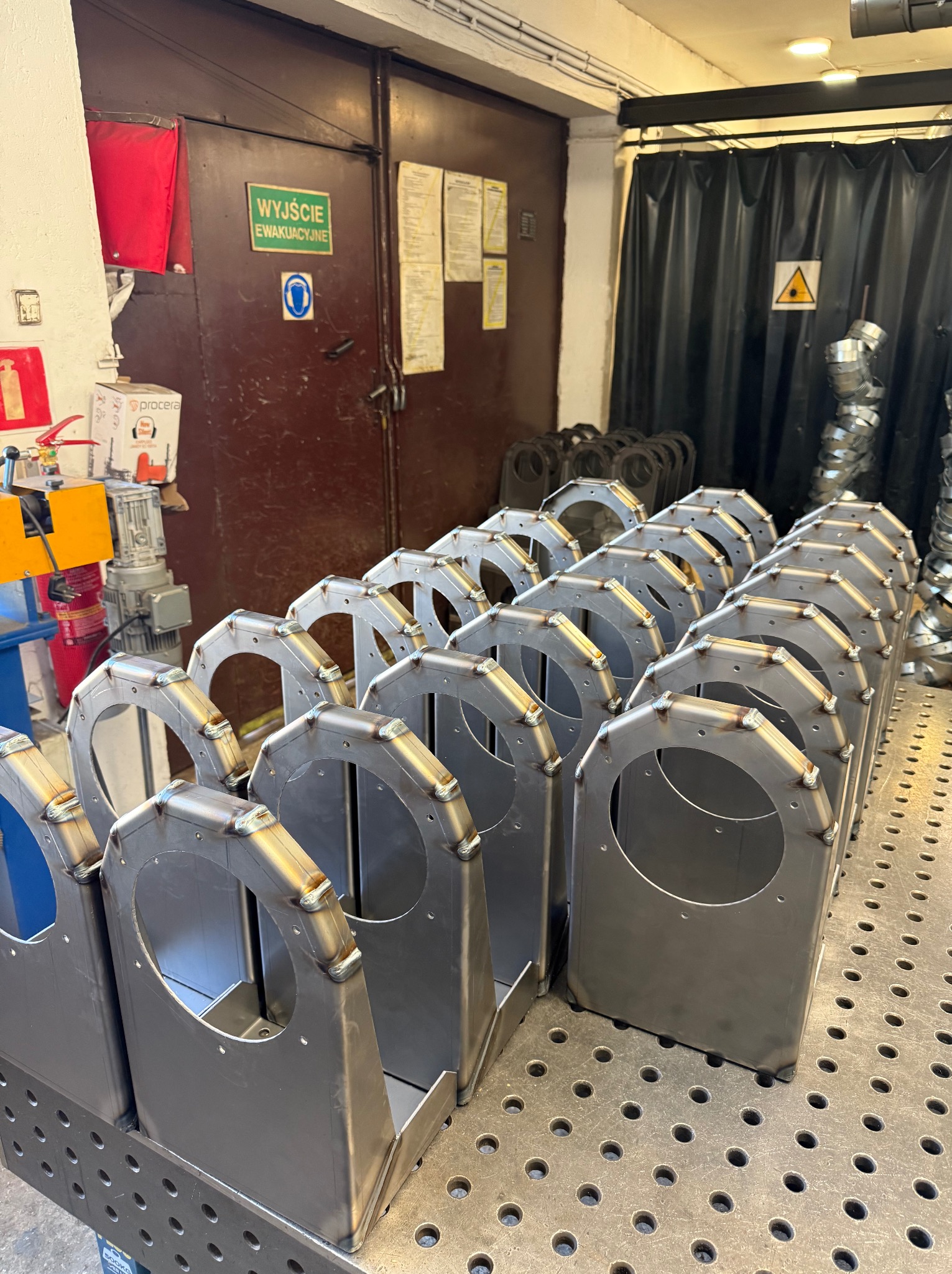 Szereg identycznych, spawanych konstrukcji metalowych z okrągłym otworem, ustawionych na perforowanym stole spawalniczym w warsztacie. Widoczne spoiny i elementy mocujące.