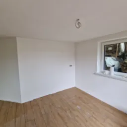 Świeżo pomalowane na biało wnętrze pokoju z drewnianą podłogą, widoczne gniazdka elektryczne i okno z widokiem na stos drewna.