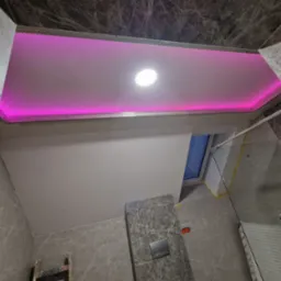 Wykończony sufit podwieszany z oświetleniem LED w kolorze różowym, widoczne płytki imitujące marmur na ścianach łazienki.