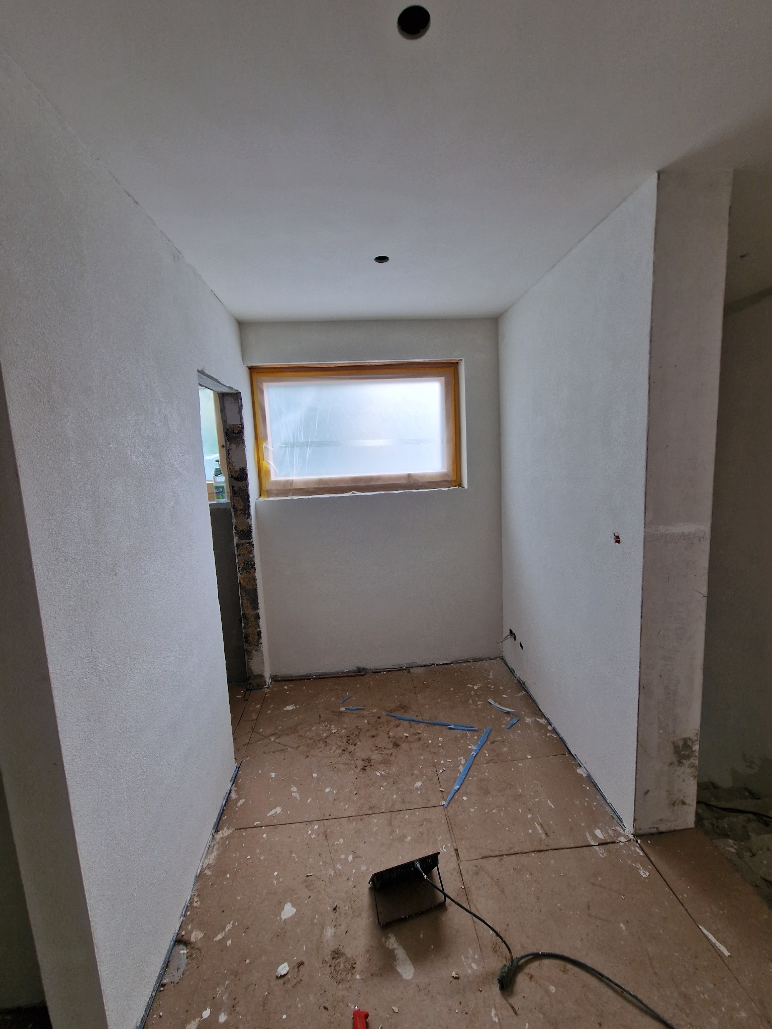 Wnętrze w trakcie prac wykończeniowych: ściany pokryte świeżą warstwą gładzi, okno zabezpieczone folią, widoczne ślady po pracach na podłodze i narzędzia.