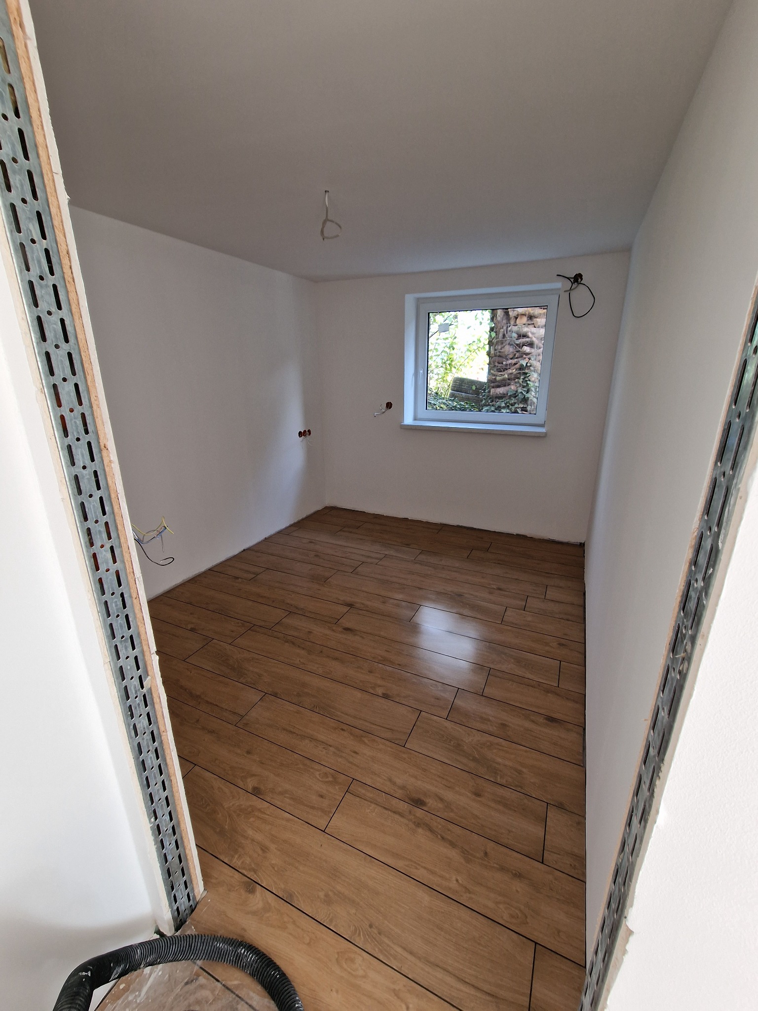 Pusty pokój po remoncie z drewnopodobną podłogą, świeżo pomalowanymi białymi ścianami i oknem z widokiem na zieleń, widoczne kable elektryczne i metalowe profile przy drzwiach.