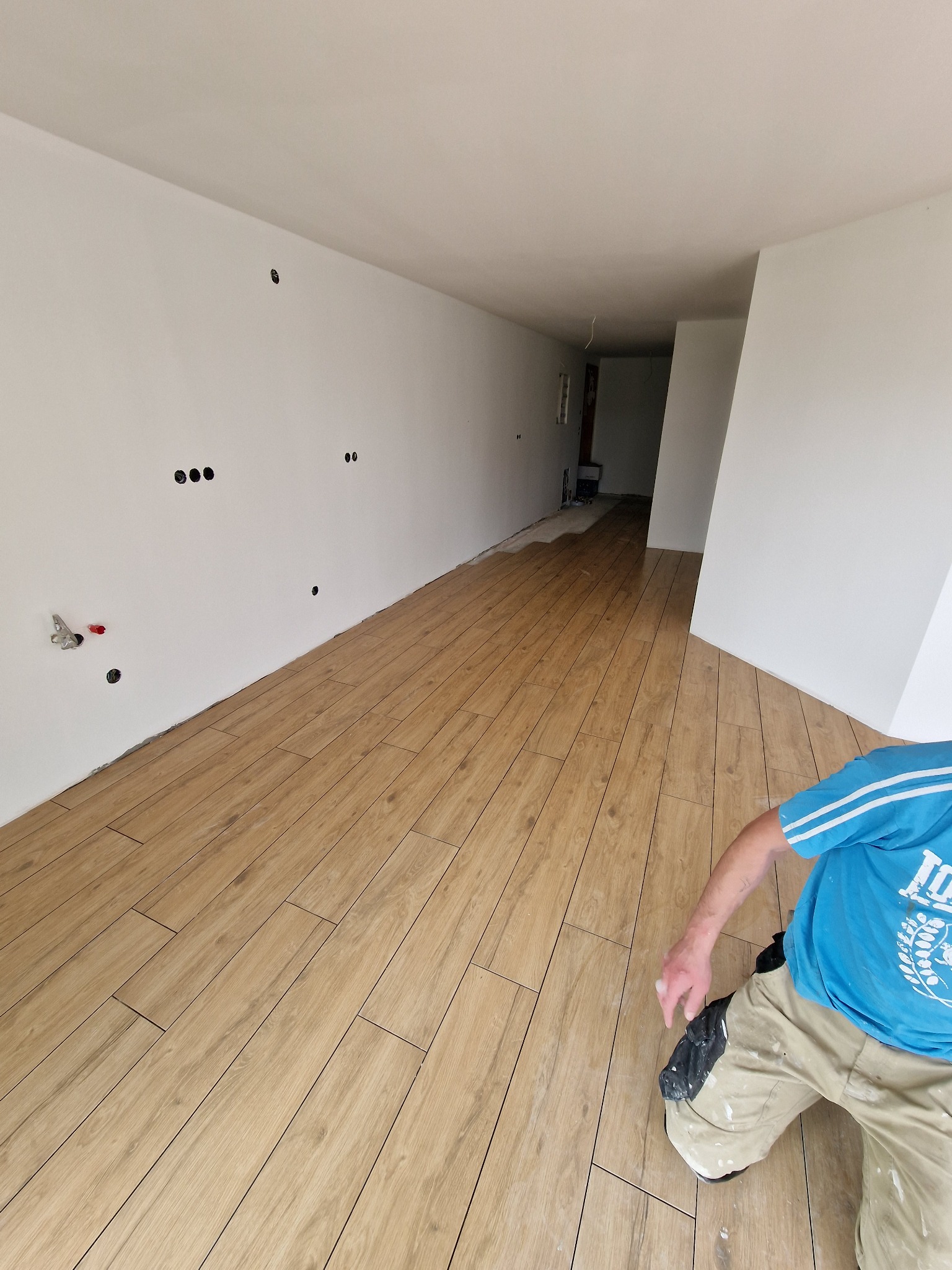 Układanie paneli podłogowych imitujących drewno w długim, pustym pomieszczeniu z białymi ścianami i przygotowanymi punktami elektrycznymi; widoczny fragment sylwetki wykonawcy w roboczym ubraniu.