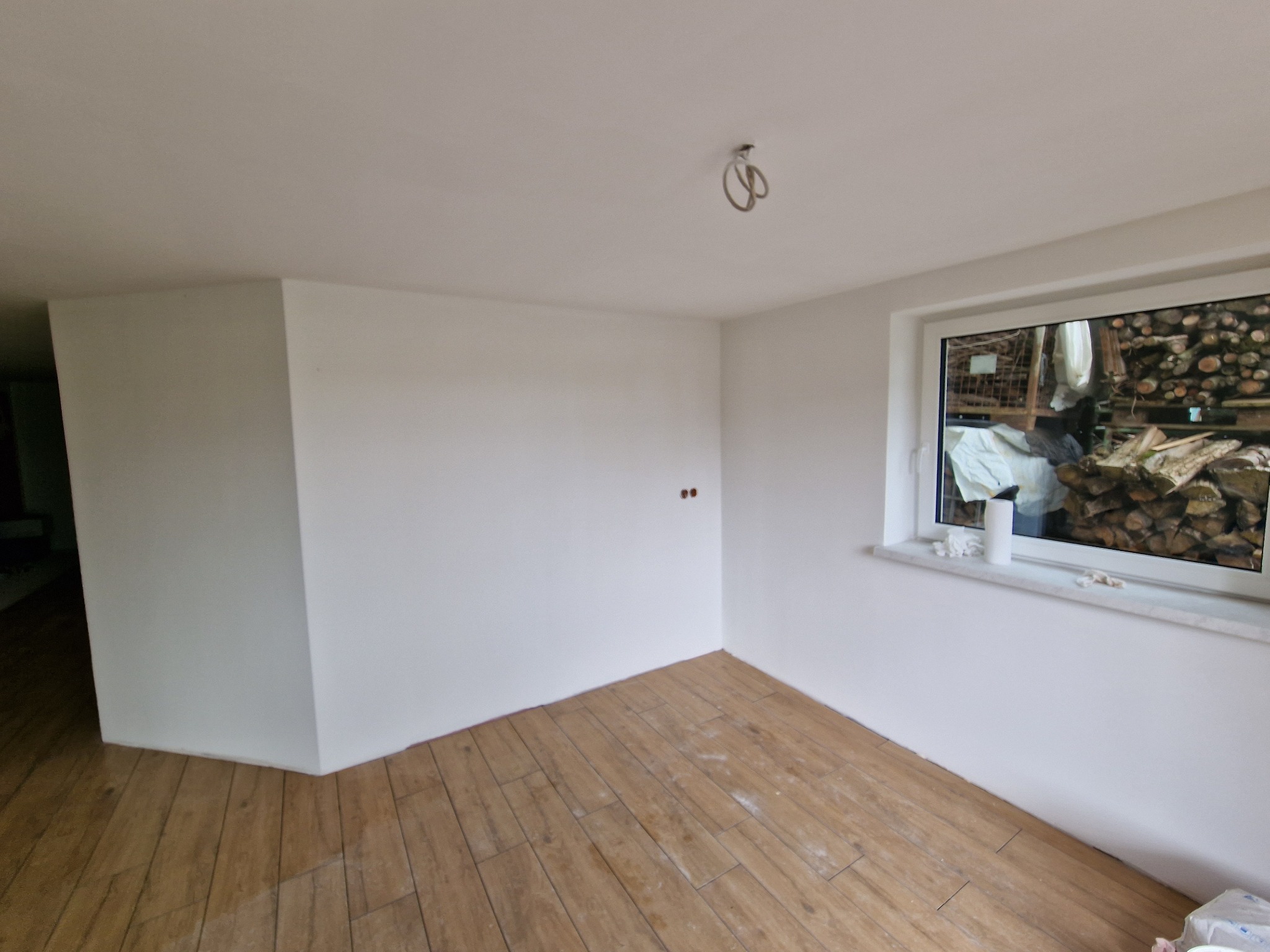 Świeżo pomalowane na biało wnętrze pokoju z drewnianą podłogą, widoczne gniazdka elektryczne i okno z widokiem na stos drewna.