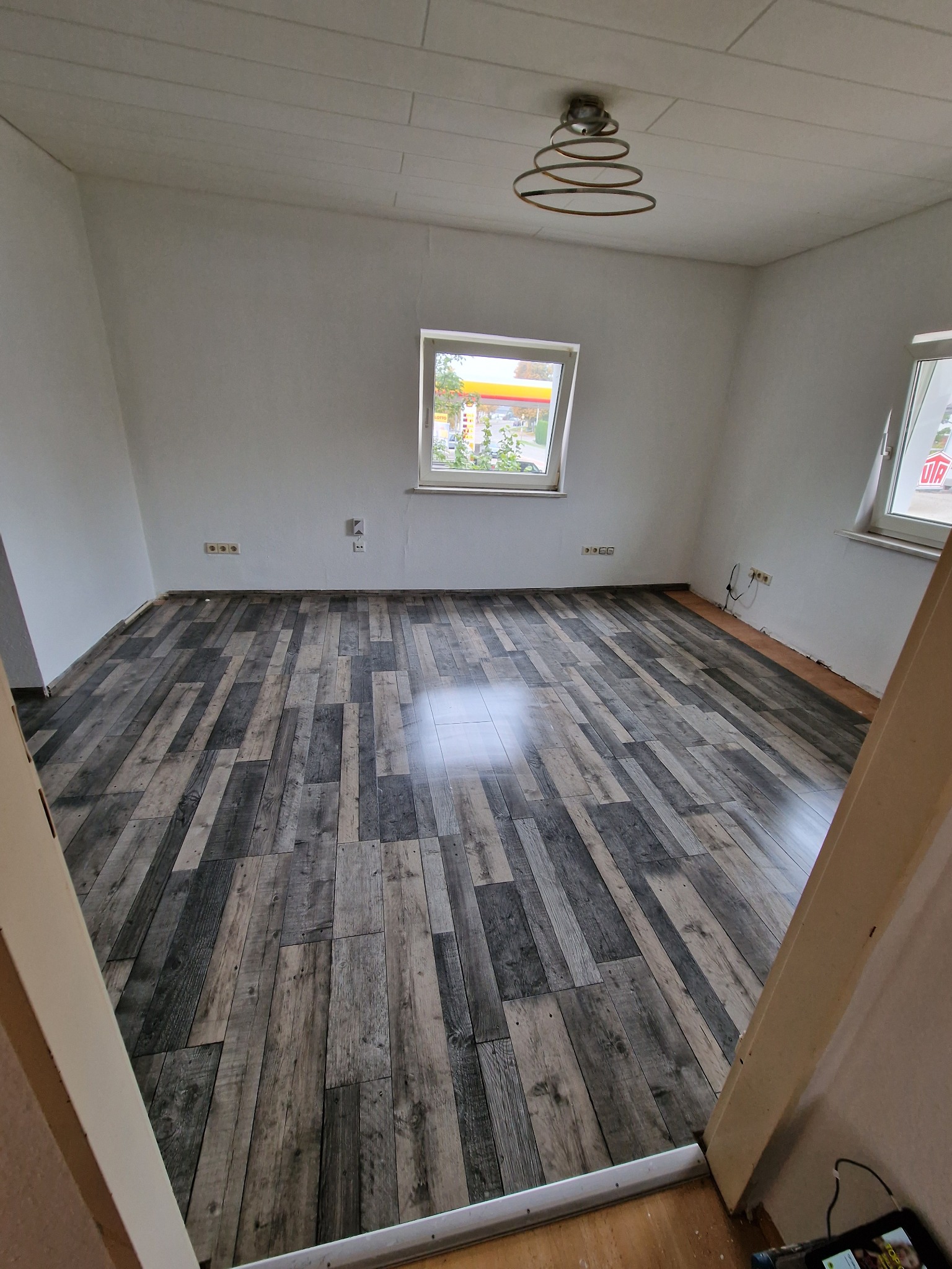 Pusty pokój po remoncie z panelami podłogowymi imitującymi szare drewno, widoczne gniazdka elektryczne i okna z widokiem na zewnątrz.