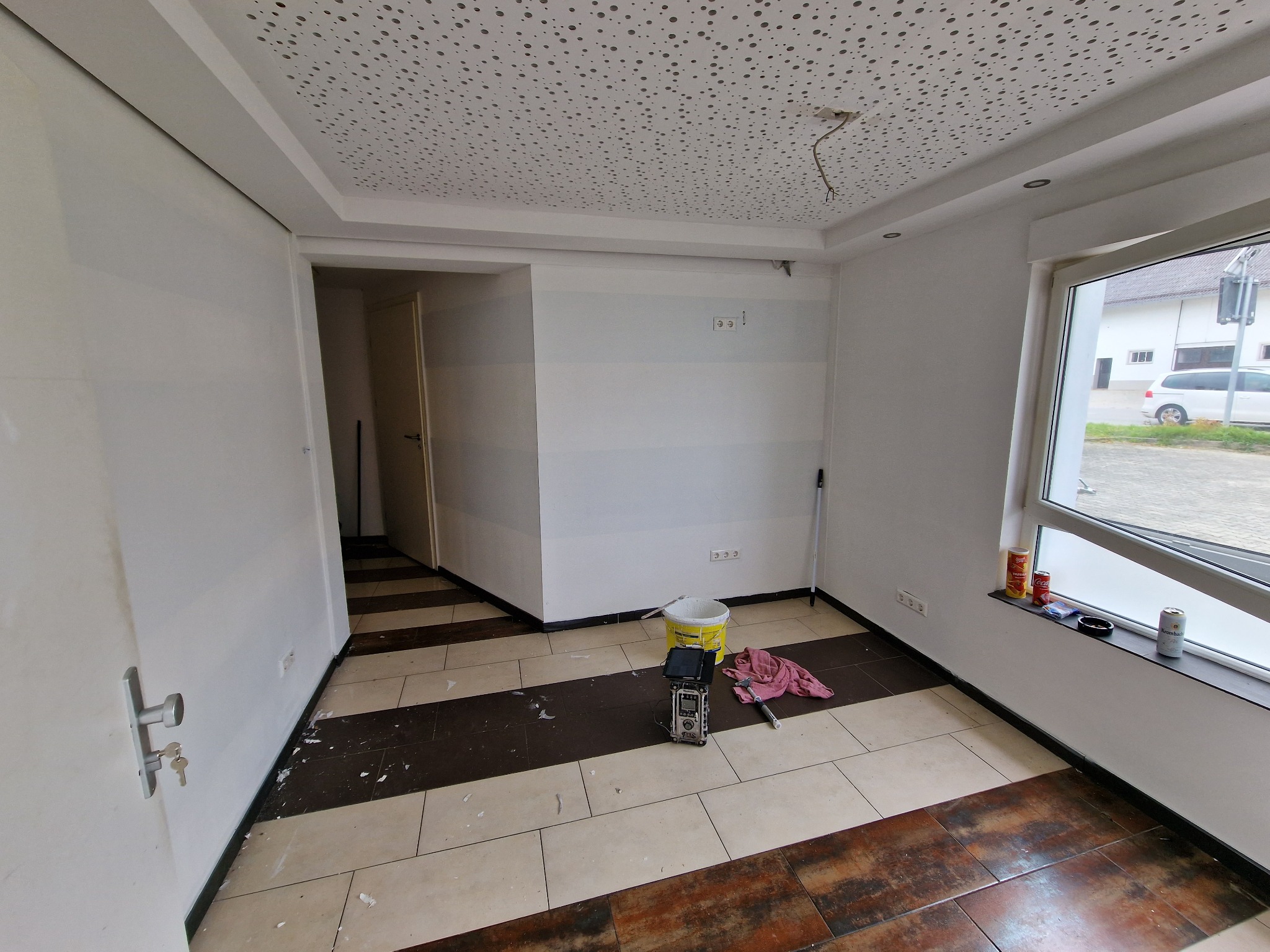 Pokój w trakcie odświeżania: widoczne białe ściany z poziomymi, szarymi pasami, perforowany sufit, wiadro z farbą, wałek malarski i resztki farby na podłodze z płytek.