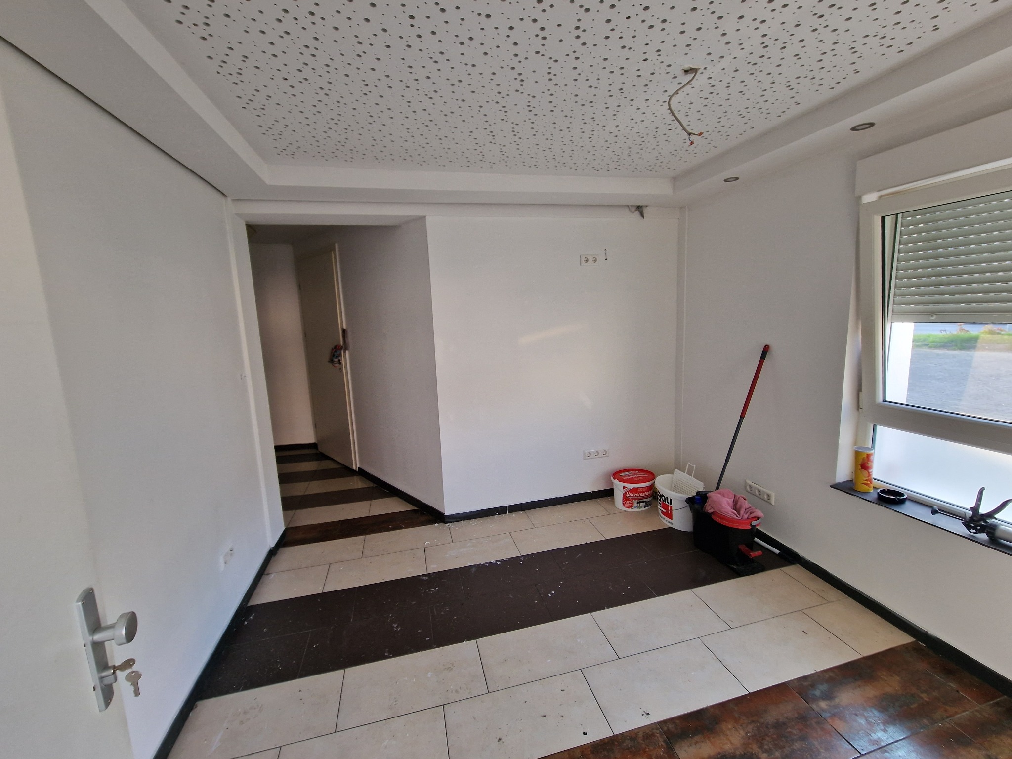 Wnętrze pokoju w trakcie odświeżania, ściany pomalowane na biało, widoczne wiadra z farbą, wałek malarski i akcesoria malarskie w rogu pomieszczenia, podłoga wyłożona płytkami w odcieniach brązu...