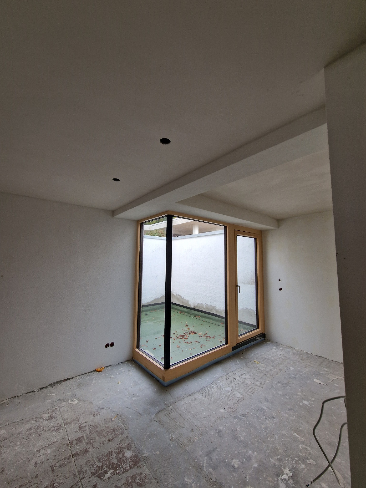 Surowe wnętrze z dużym narożnym oknem z drewnianą ramą, ściany w trakcie przygotowania do malowania, widoczne niedoskonałości i ślady po pracach wykończeniowych, podłoga z resztkami kleju lub zaprawy.