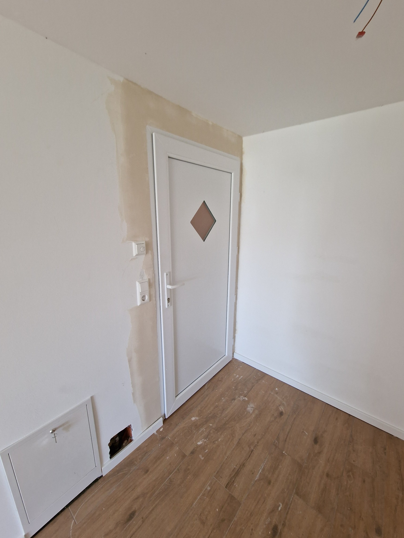 Wnętrze pokoju w trakcie prac wykończeniowych: widoczne fragmenty ściany z pozostałościami po szpachlowaniu, białe drzwi z brązowym rombem, drewnopodobna podłoga oraz otwór w ścianie obok skrzynki...