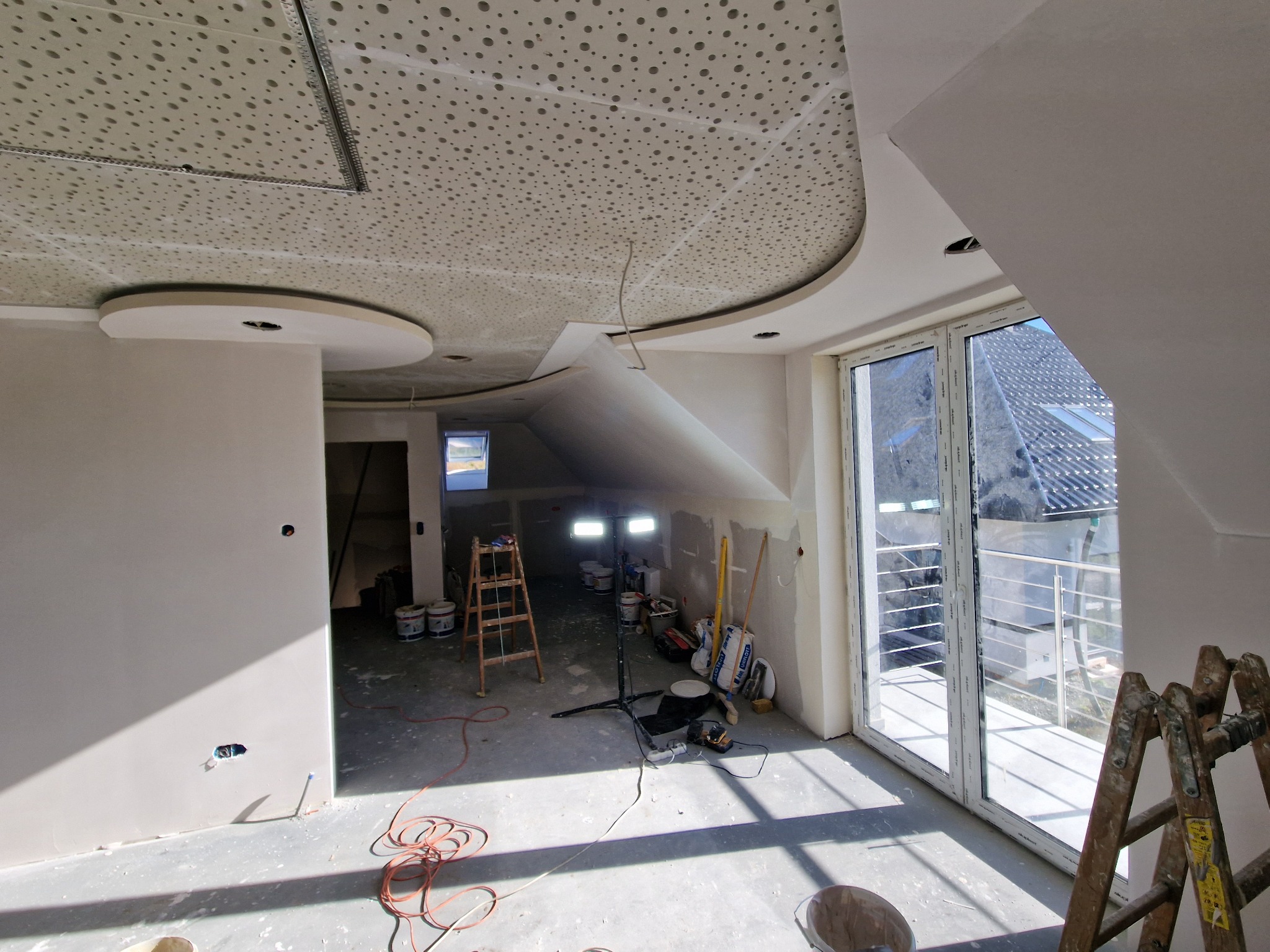 Wnętrze w trakcie wykańczania: szpachlowane ściany, sufit z perforacją, drabiny, narzędzia i worki z materiałami budowlanymi, widoczne przez okno zadaszenie balkonu sąsiedniego budynku.