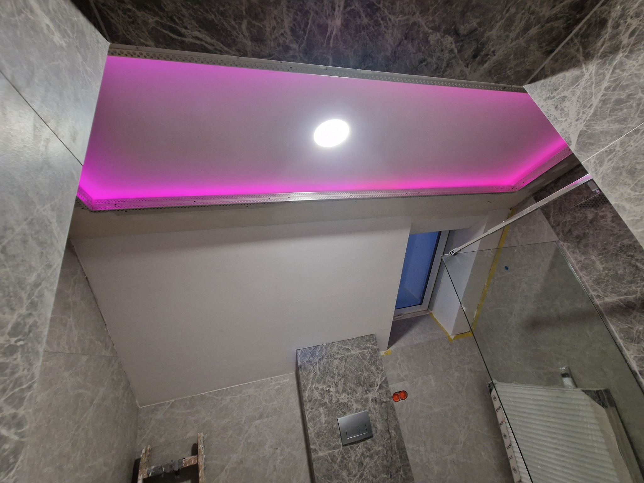Wykończony sufit podwieszany z oświetleniem LED w kolorze różowym, widoczne płytki imitujące marmur na ścianach łazienki.