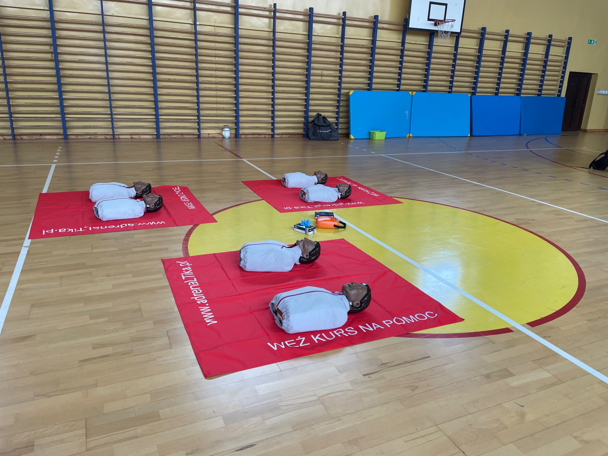 Manekiny do ćwiczeń resuscytacji na czerwonych matach z napisem na podłodze hali sportowej, sprzęt do nauki pierwszej pomocy w tle.