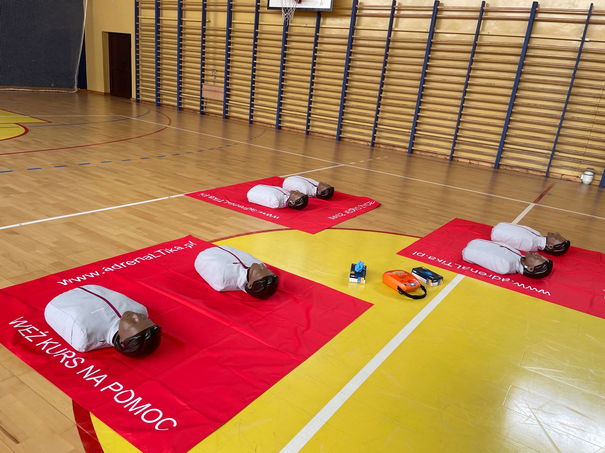 Sala gimnastyczna z czterema manekinami do resuscytacji na czerwonych matach z napisem 'Weź kurs na pomoc' i adresem strony internetowej, obok leżą akcesoria treningowe.