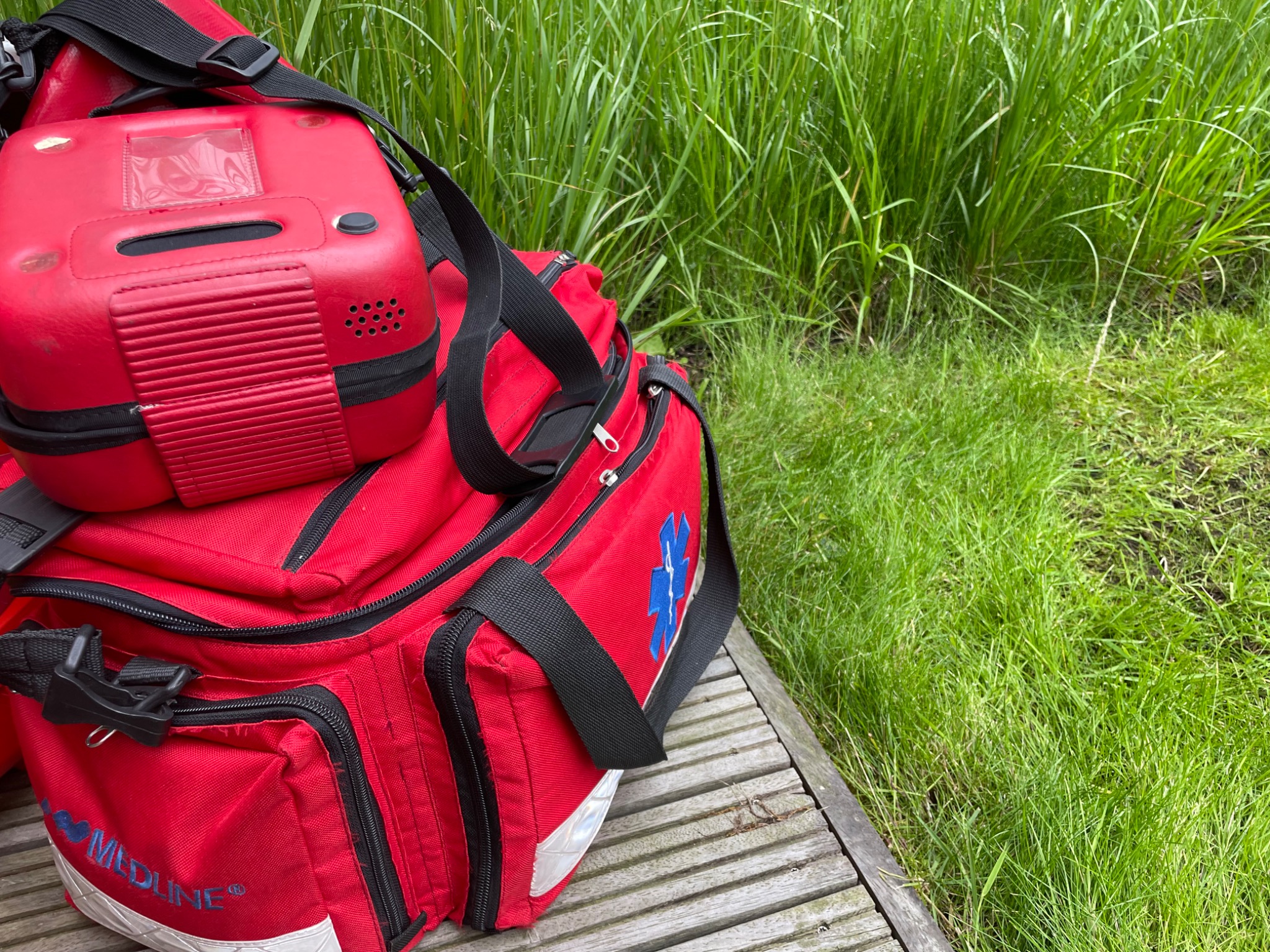 Czerwona torba medyczna z symbolem niebieskiej gwiazdy życia i defibrylator na drewnianym pomoście, w tle trawa.