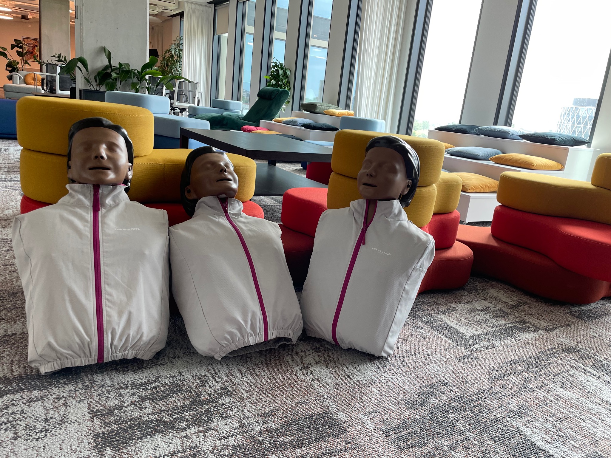 Trzy manekiny Little Anne QCPR ubrane w białe kurtki z różowymi zamkami, ustawione w nowoczesnym biurze z kolorowymi pufami i dużymi oknami.