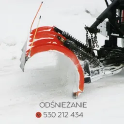 Czerwony pług śnieżny zamontowany na czarnym pojeździe odśnieża drogę pokrytą śniegiem, widoczne łańcuchy i sprężyny mocujące pług.