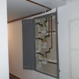 Otwarta szafka z trzema licznikami gazu, rurami miedzianymi i żółtymi zaworami w korytarzu, widoczne rury prowadzone pod sufitem.