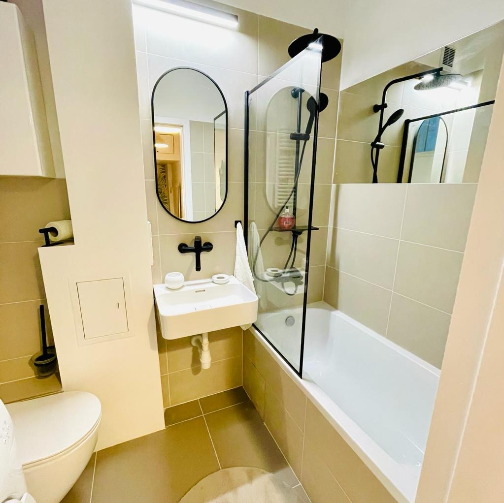 Nowoczesna łazienka w beżach z czarnymi akcentami: owalne lustro, bateria umywalkowa w kształcie krzyża, kabina prysznicowa z czarną ramą, płytki ceramiczne i biała wanna.