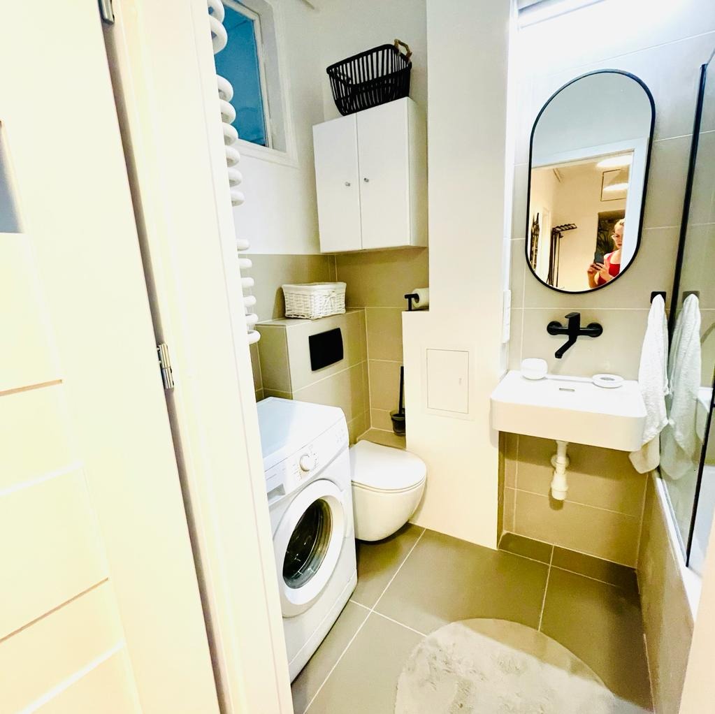 Nowoczesna, funkcjonalna łazienka z pralką, umywalką nablatową z czarną armaturą, owalnym lustrem i szarą glazurą, doświetlona naturalnym światłem z okna.