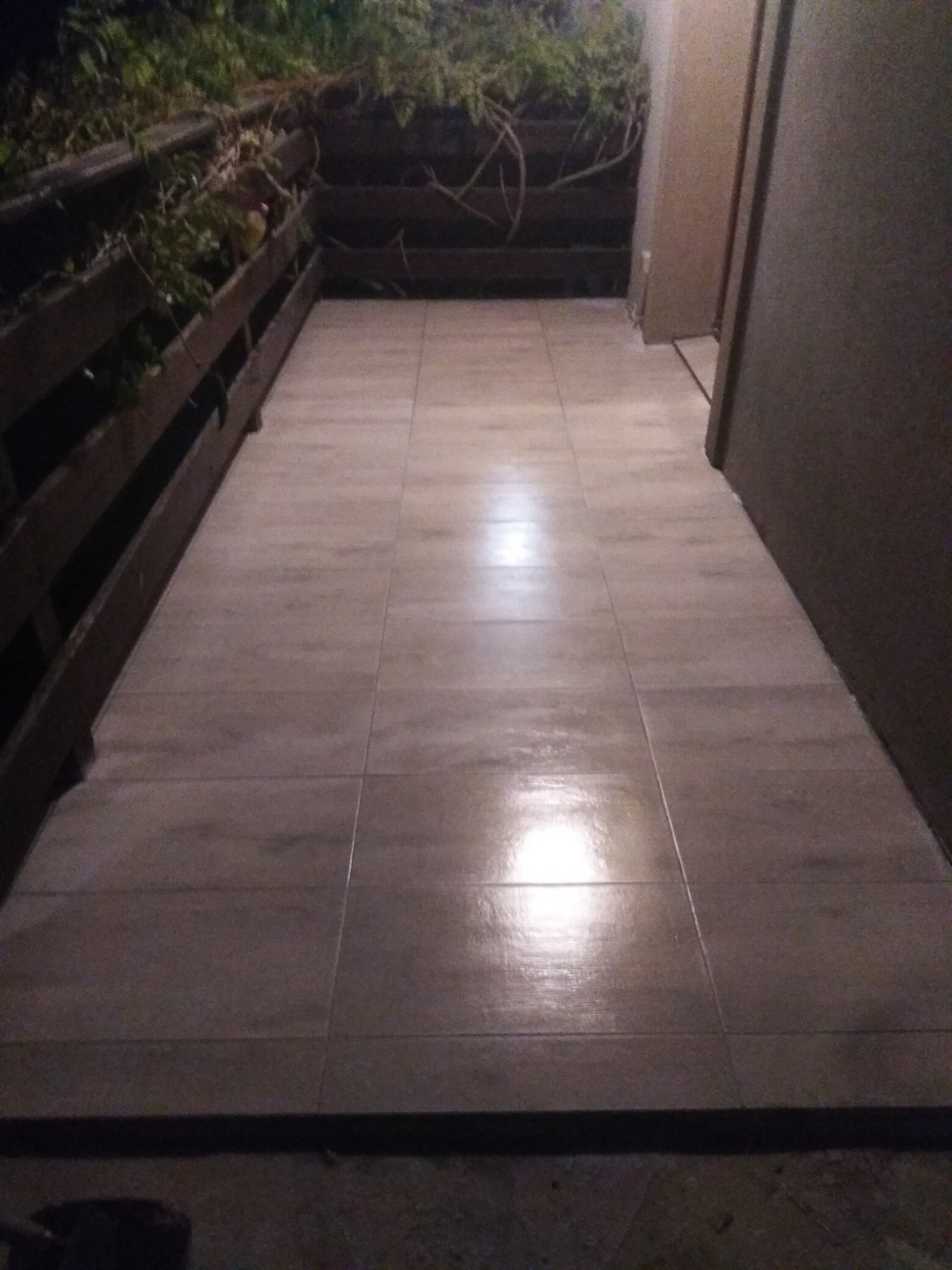 Wyłożona płytkami podłoga balkonu z drewnianą balustradą i roślinnością w tle, oświetlona sztucznym światłem.