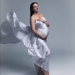 Olena Ekiert - Sesja ciążowa w Warszawie: Kobieta w zwiewnej, białej tkaninie, pozuje z zamkniętymi oczami i dłonią na brzuchu, tkanina faluje w powietrzu.