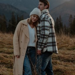 Olena Ekiert - Zakochana para w jesiennej scenerii górskiej: kobieta w beżowym płaszczu i czapce opiera głowę na ramieniu mężczyzny w kraciastej koszuli, oboje ubrani w jeansy, na tle zalesionych szczytów...