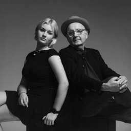 Olena Ekiert - Czarno-białe portretowe ujęcie kobiety z blond włosami i mężczyzny w kapeluszu i okularach, siedzących obok siebie na ciemnym tle.