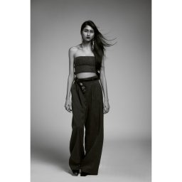 Olena Ekiert - Pełna sylwetka kobiety w modnym stroju: top typu tuba, szerokie spodnie z paskiem i długie kolczyki, włosy rozwiane wiatrem na szarym tle.