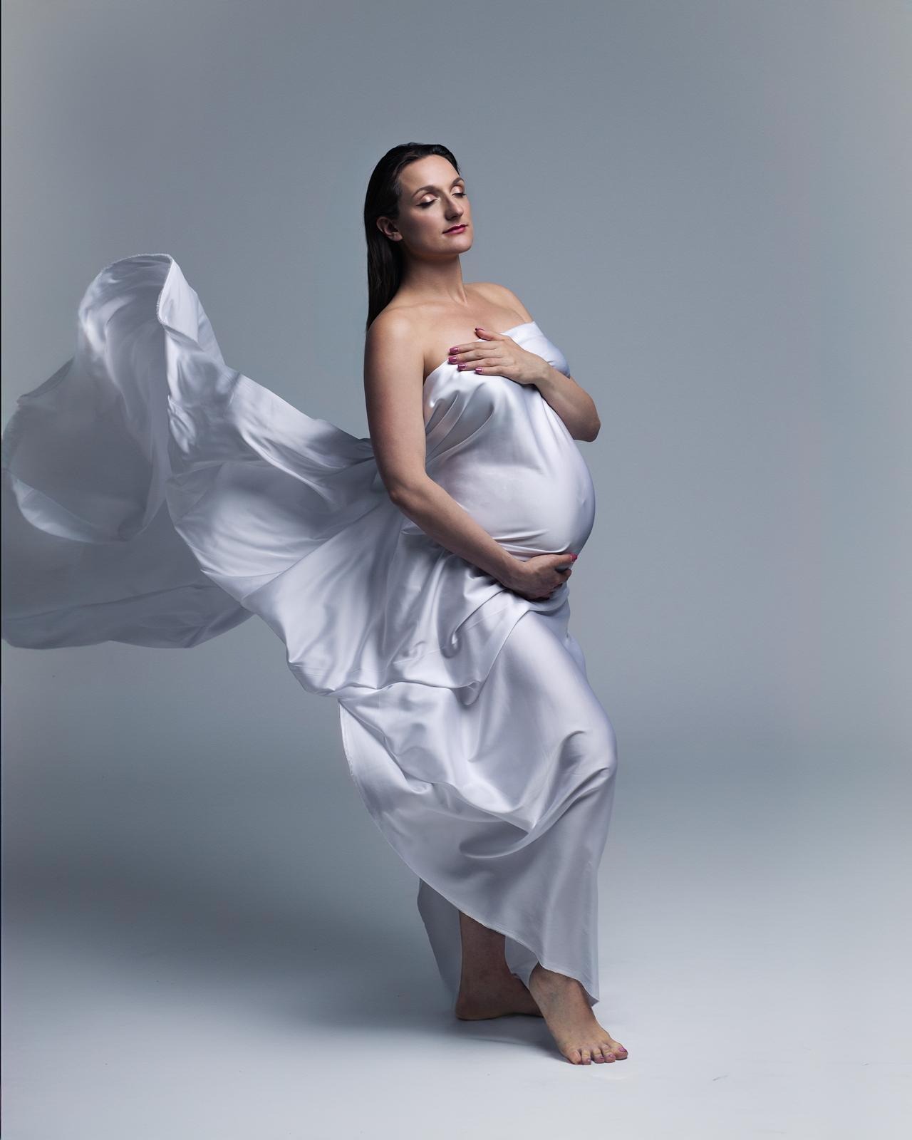 Sesja ciążowa w Warszawie: Kobieta w zwiewnej, białej tkaninie, pozuje z zamkniętymi oczami i dłonią na brzuchu, tkanina faluje w powietrzu.