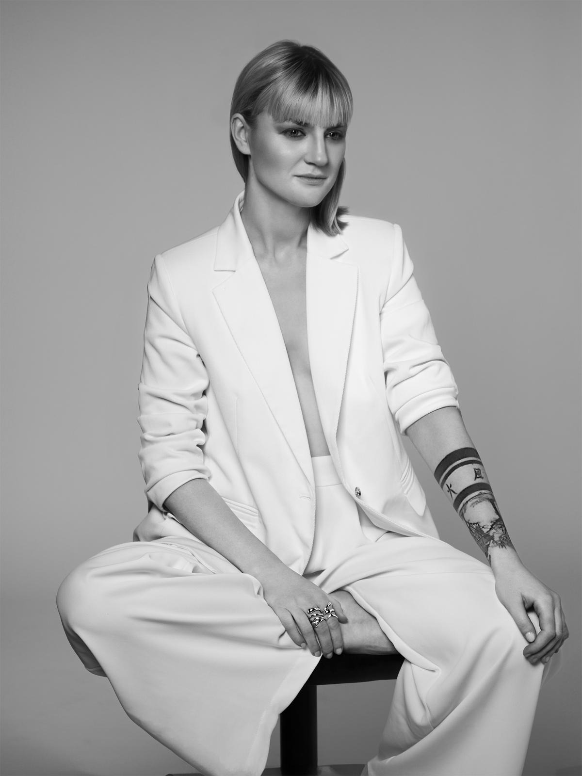 Czarno-białe portretowe zdjęcie kobiety w białym garniturze, siedzącej na stołku, z widocznym tatuażem na ramieniu i dużym pierścieniem na palcu.