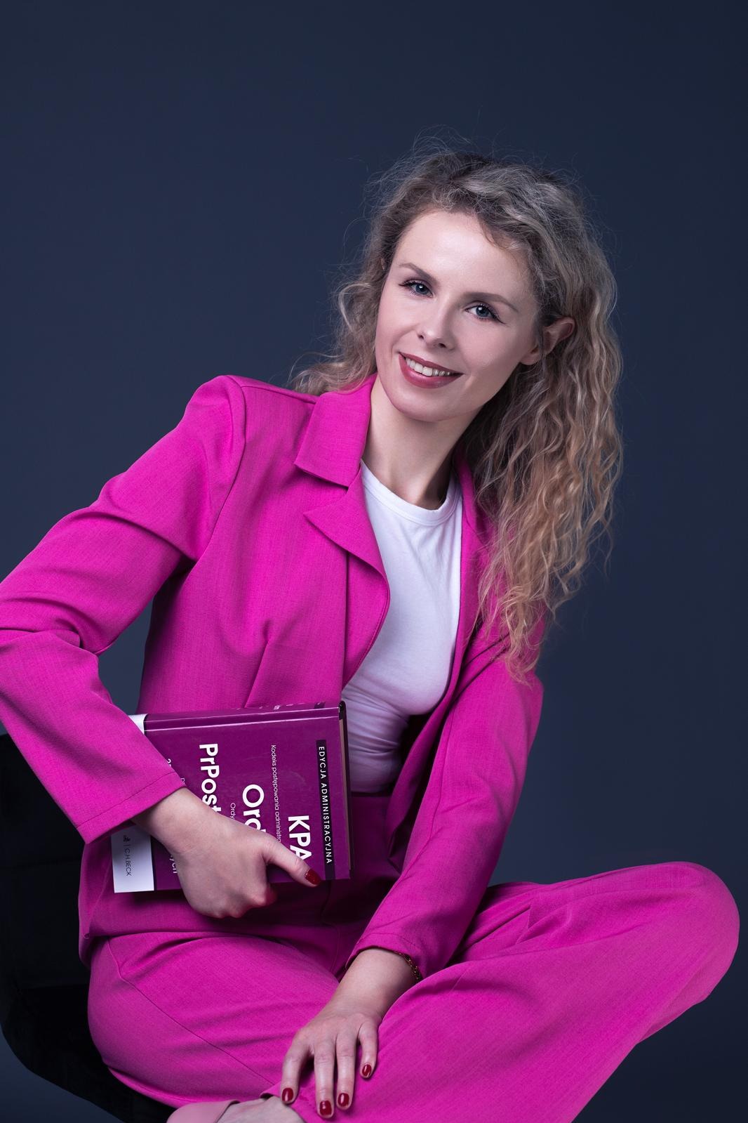 Uśmiechnięta blondynka w różowym garniturze, siedząca na krześle na ciemnym tle, trzymająca w dłoniach książkę z widocznym tytułem.