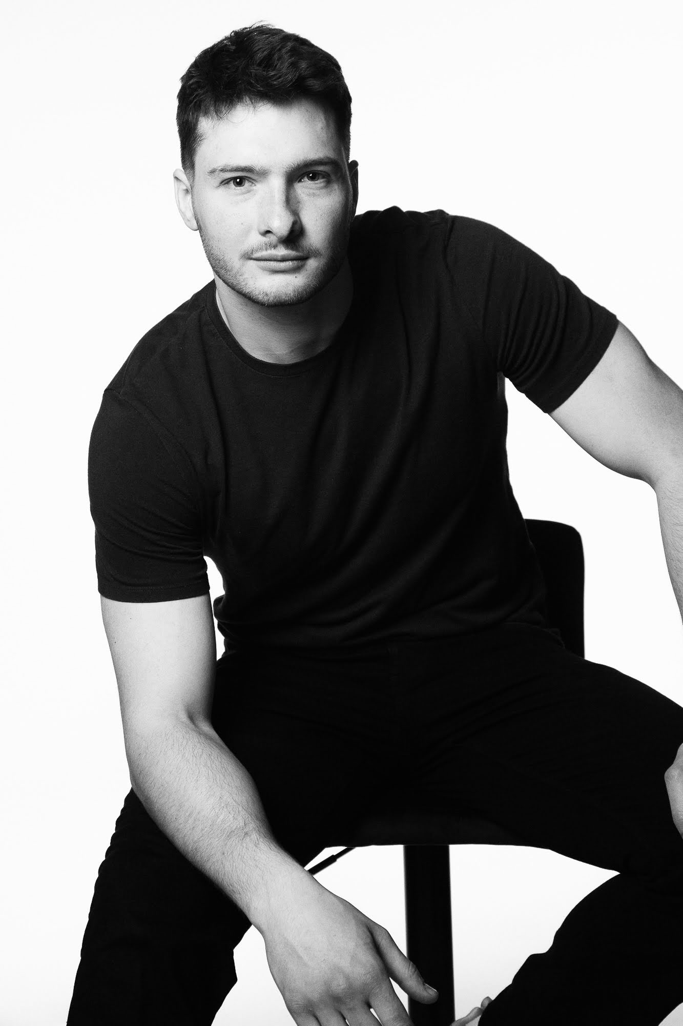 Portret młodego mężczyzny w czarnym t-shircie i spodniach, siedzącego na krześle na białym tle, ujęcie w czerni i bieli.