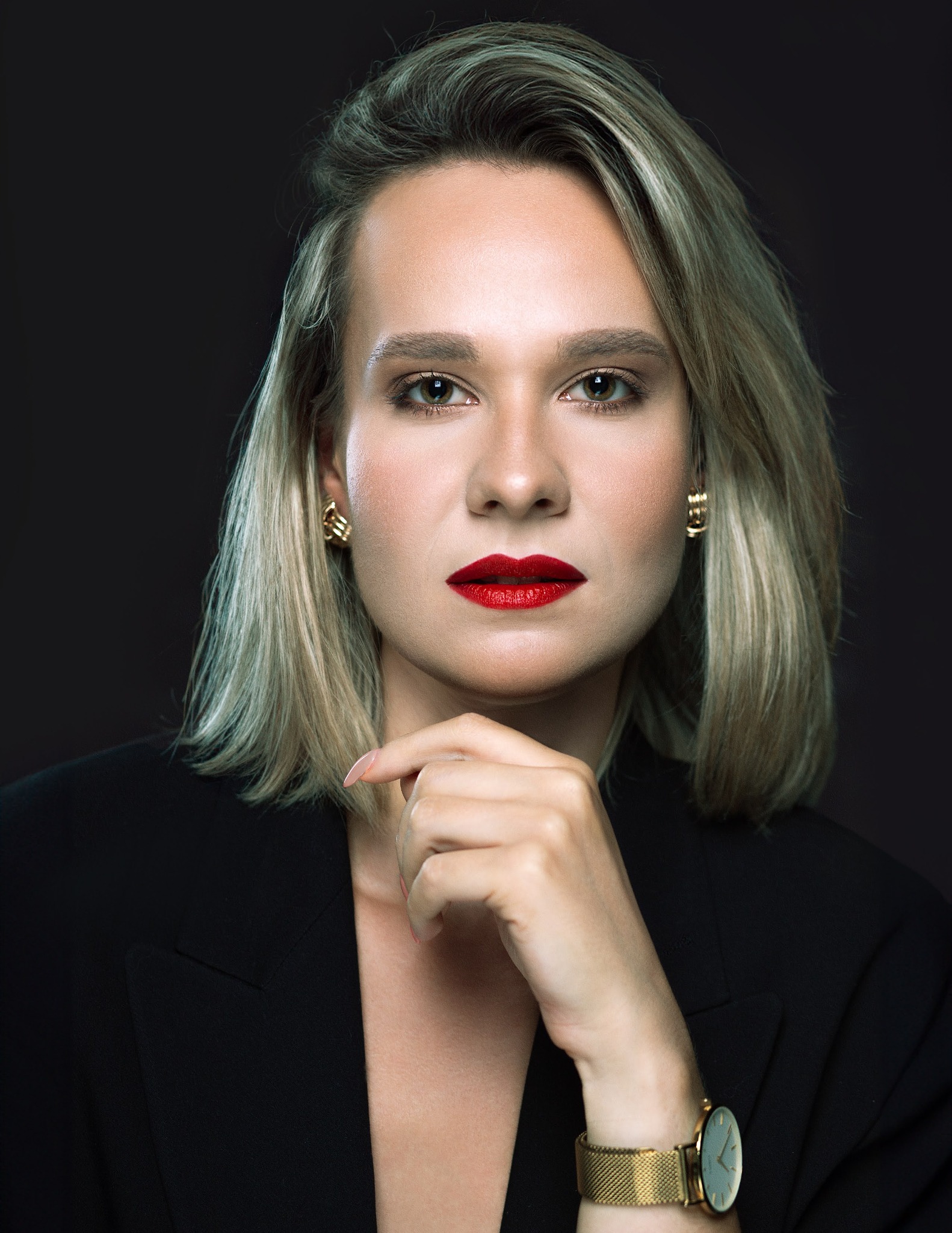 Portret kobiety w czarnej marynarce, z blond włosami, czerwonymi ustami i złotym zegarkiem, pozującej na ciemnym tle.