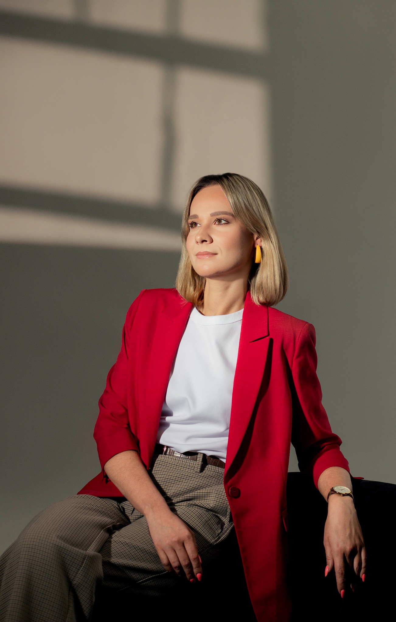 Portret kobiety w czerwonej marynarce, białej koszulce i spodniach w kratę, siedzącej na czarnej sofie, z żółtymi kolczykami i zegarkiem na ręce, z delikatnym uśmiechem i wzrokiem skierowanym...