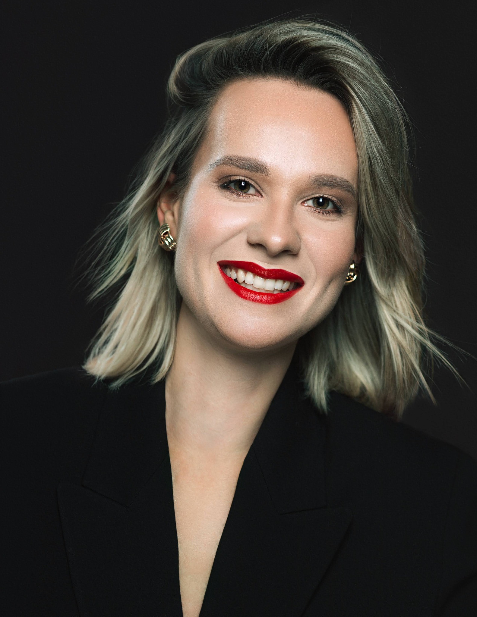 Portret uśmiechniętej kobiety z blond włosami, w czarnej marynarce, z intensywnie czerwoną szminką i złotymi kolczykami na ciemnym tle.