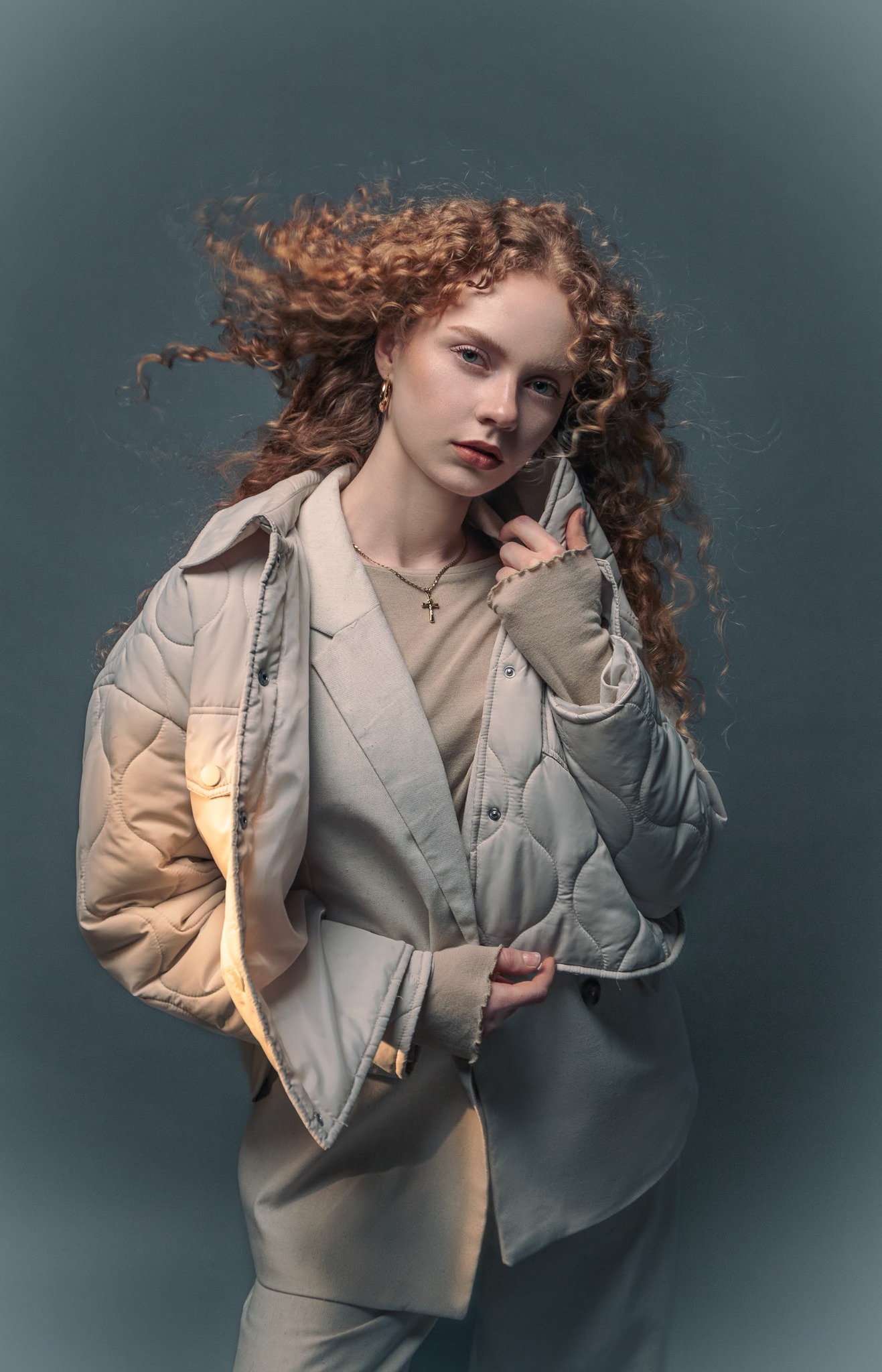 Portret młodej kobiety z burzą rudych loków, ubrana w beżową kurtkę i bluzkę, z delikatnym makijażem i złotym naszyjnikiem z krzyżykiem, na jednolitym szarym tle.