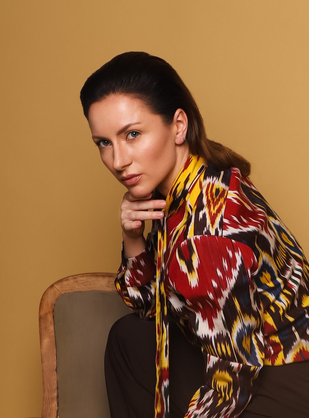 Portret kobiety z niebieskimi oczami, opierającej brodę na dłoni, ubrana w wzorzystą, kolorową bluzkę siedzącej na krześle na jednolitym, ciepłym tle.