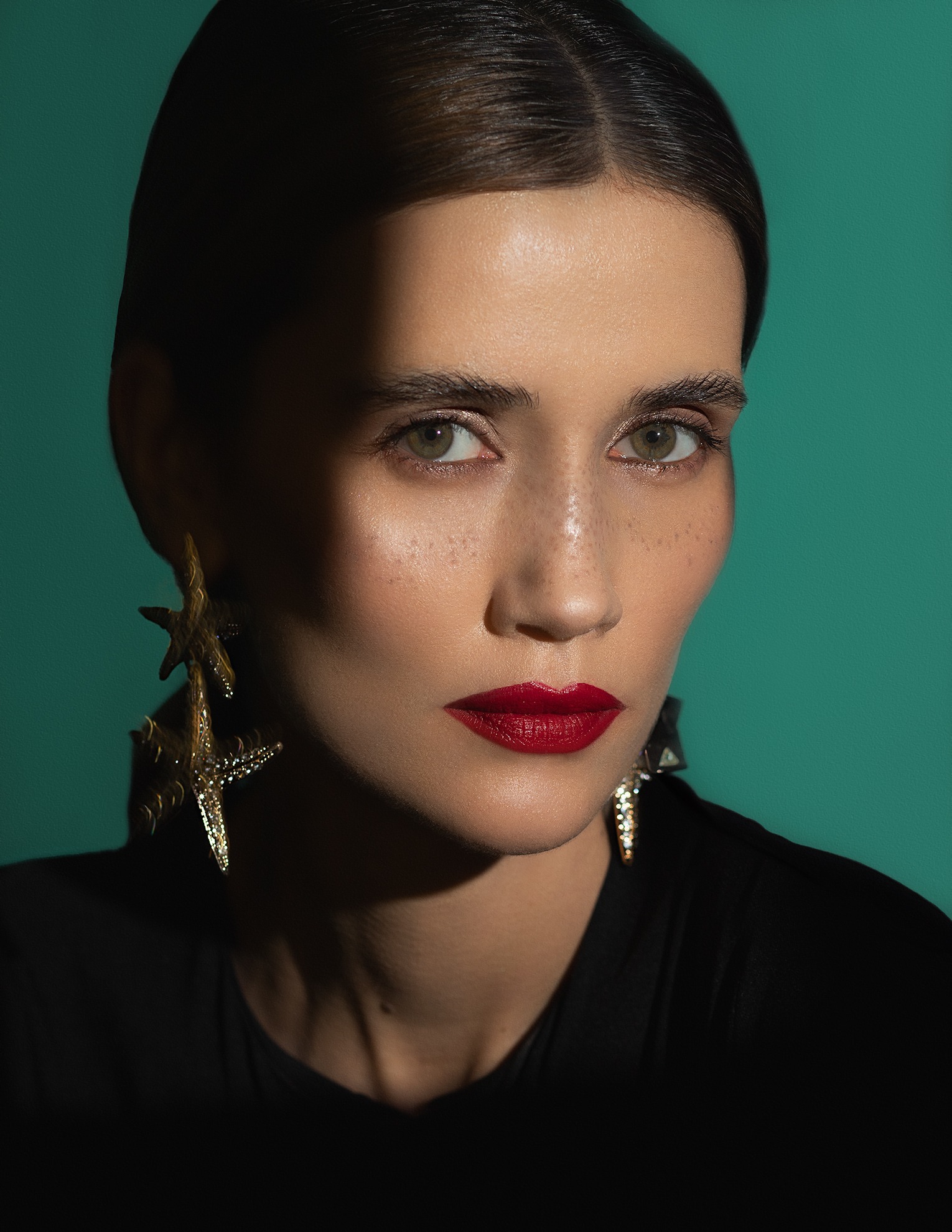 Portret kobiety z intensywnie czerwoną szminką, geometrycznym cieniem na twarzy i dużymi kolczykami w kształcie gwiazd na tle turkusowego ekranu.