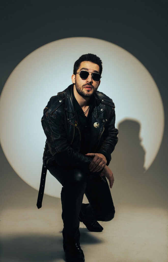 Mężczyzna w czarnej skórzanej kurtce i okularach przeciwsłonecznych pozuje w klęczącej pozycji na tle okrągłego światła.