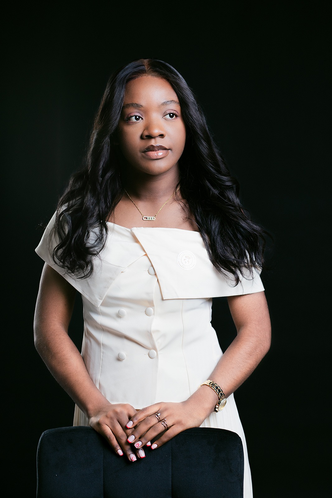 Portret młodej kobiety z ciemnymi włosami w kremowej sukience, pozującej na czarnym tle, z dłońmi opartymi na krześle, widoczna biżuteria: naszyjnik, pierścionki i zegarek.